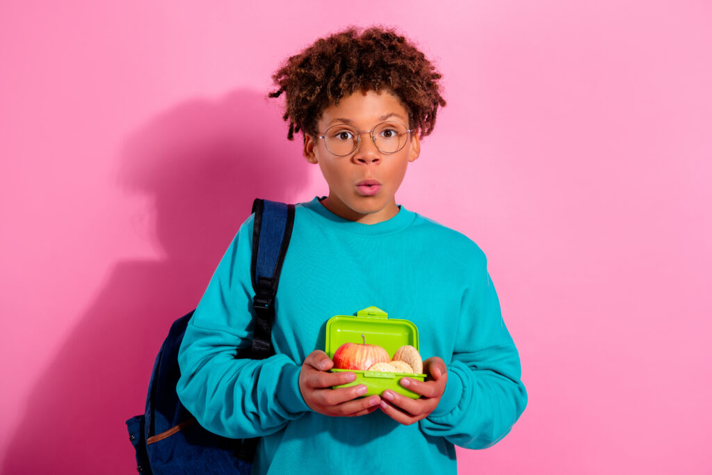 The Parent Communication Gap in School Nutrition: Why Great Programs Go Unnoticed Surprised school student holding a green lunchbox with apple and snacks while wearing backpack against pink background